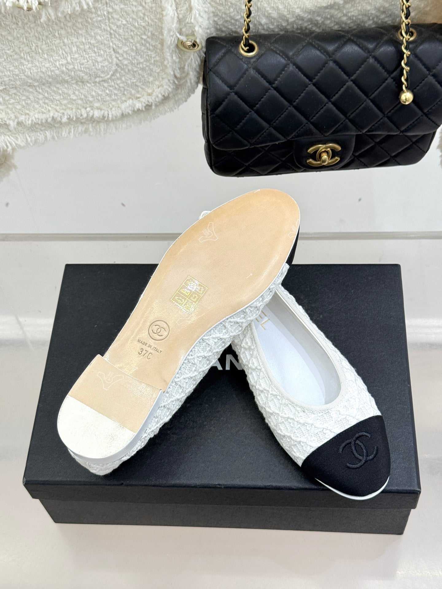 White lace ballet flats with black toe caps and CC emblem, with sole visible on shoebox