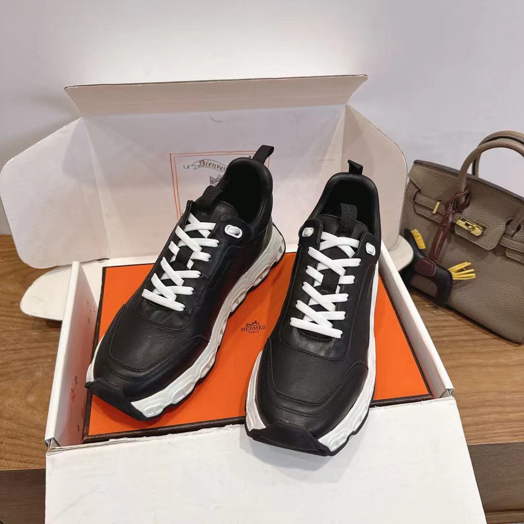 Black unisex sneakers in open shoe box, white laces, thick white sole.