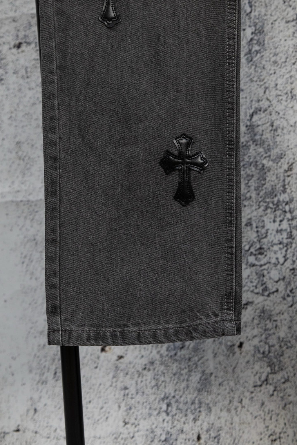 Dark gray denim pants hem with black cross appliqué near the cuff