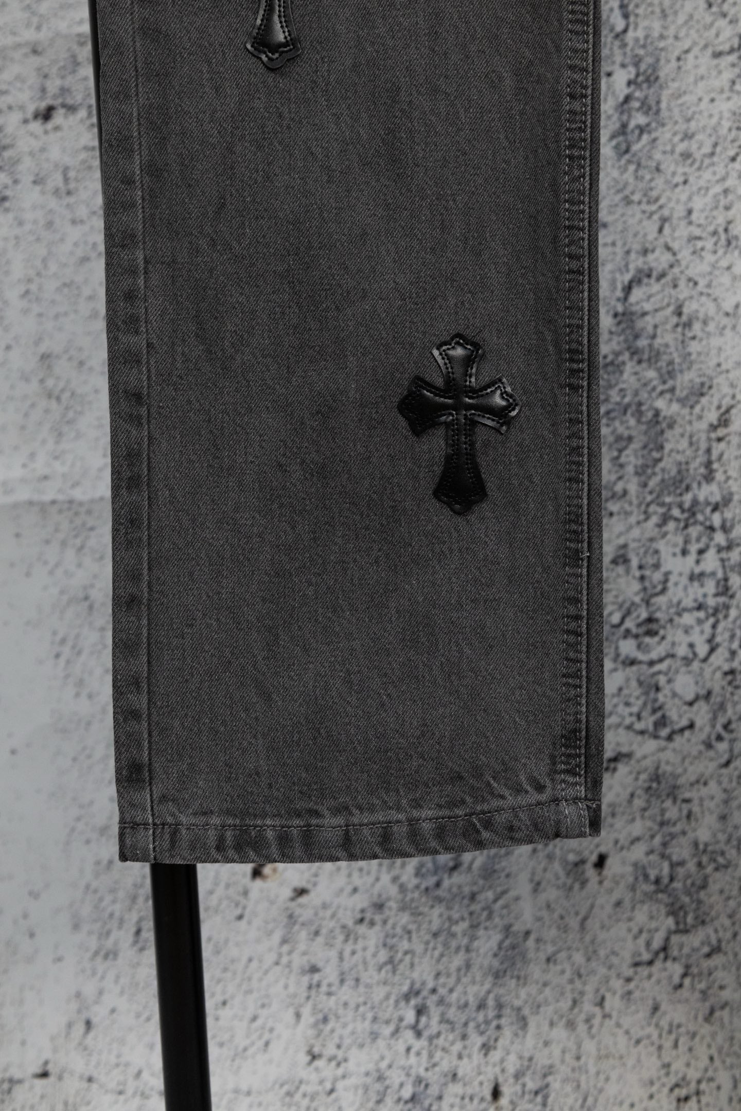 Dark gray denim pants hem with black cross appliqué near the cuff