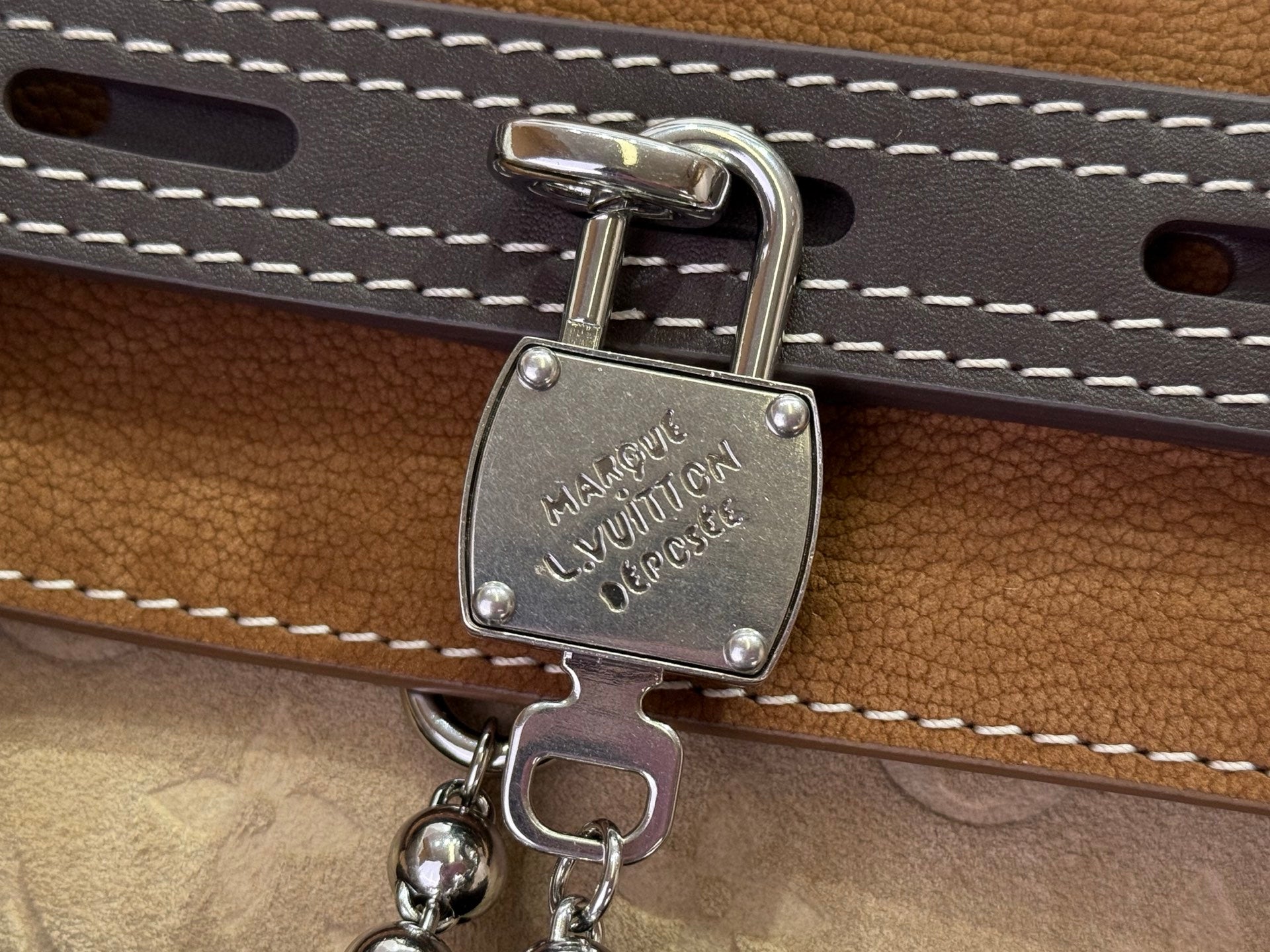 Louis Vuitton engraved metal lock charm on tan leather strap with white stitching.