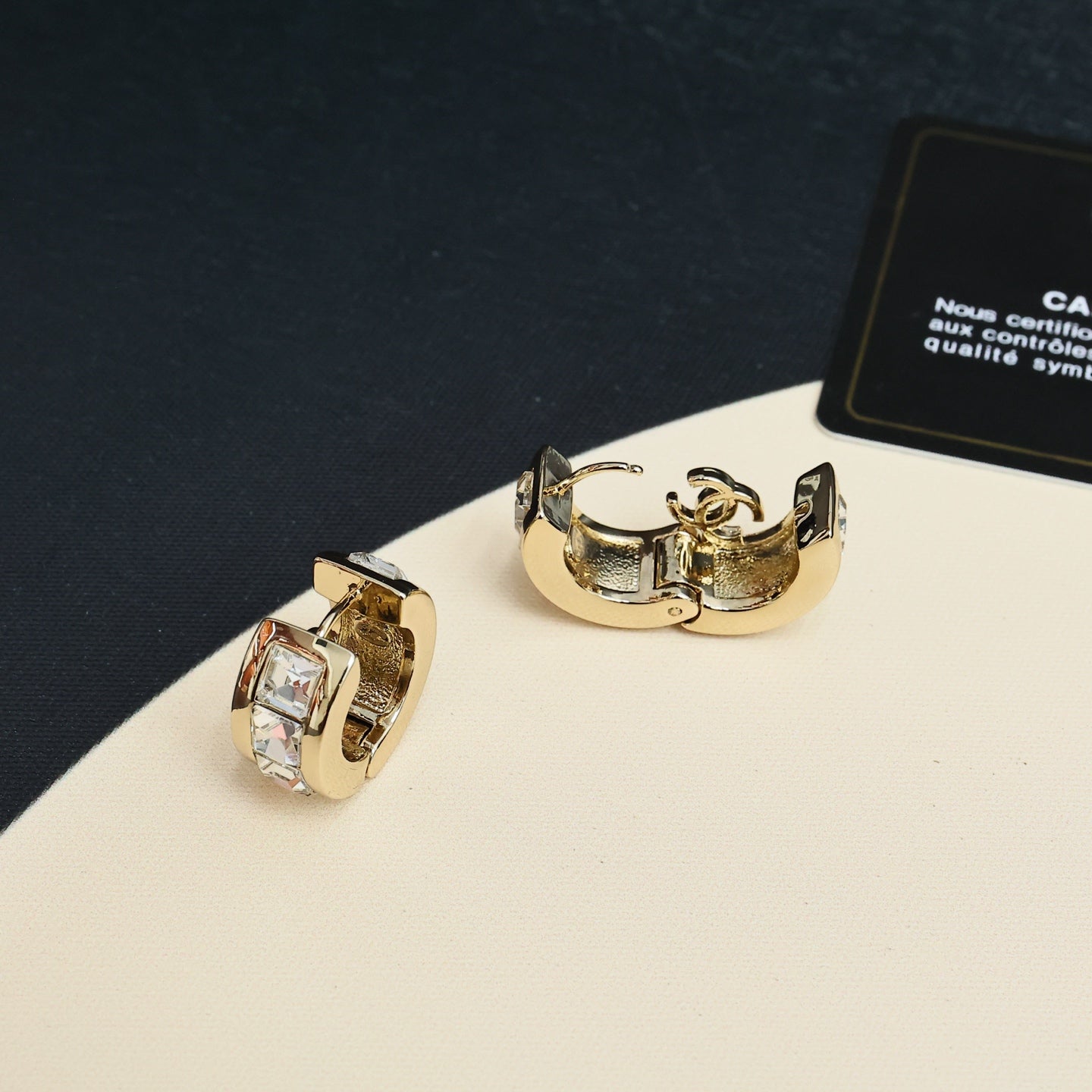 Gold-tone hoop earrings with clear rhinestone center and logo emblem, angled view