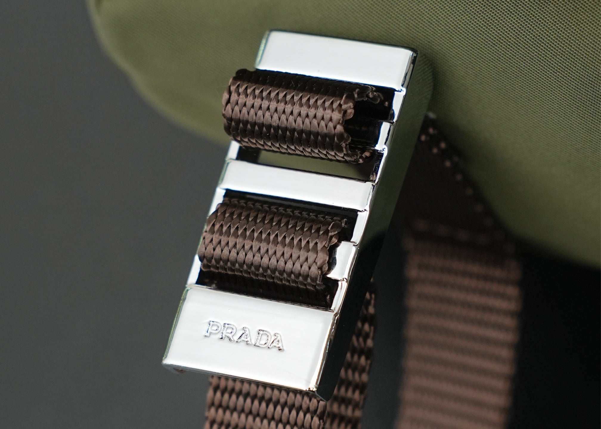 Prada-engraved metal buckle with brown woven webbing strap on green bag.