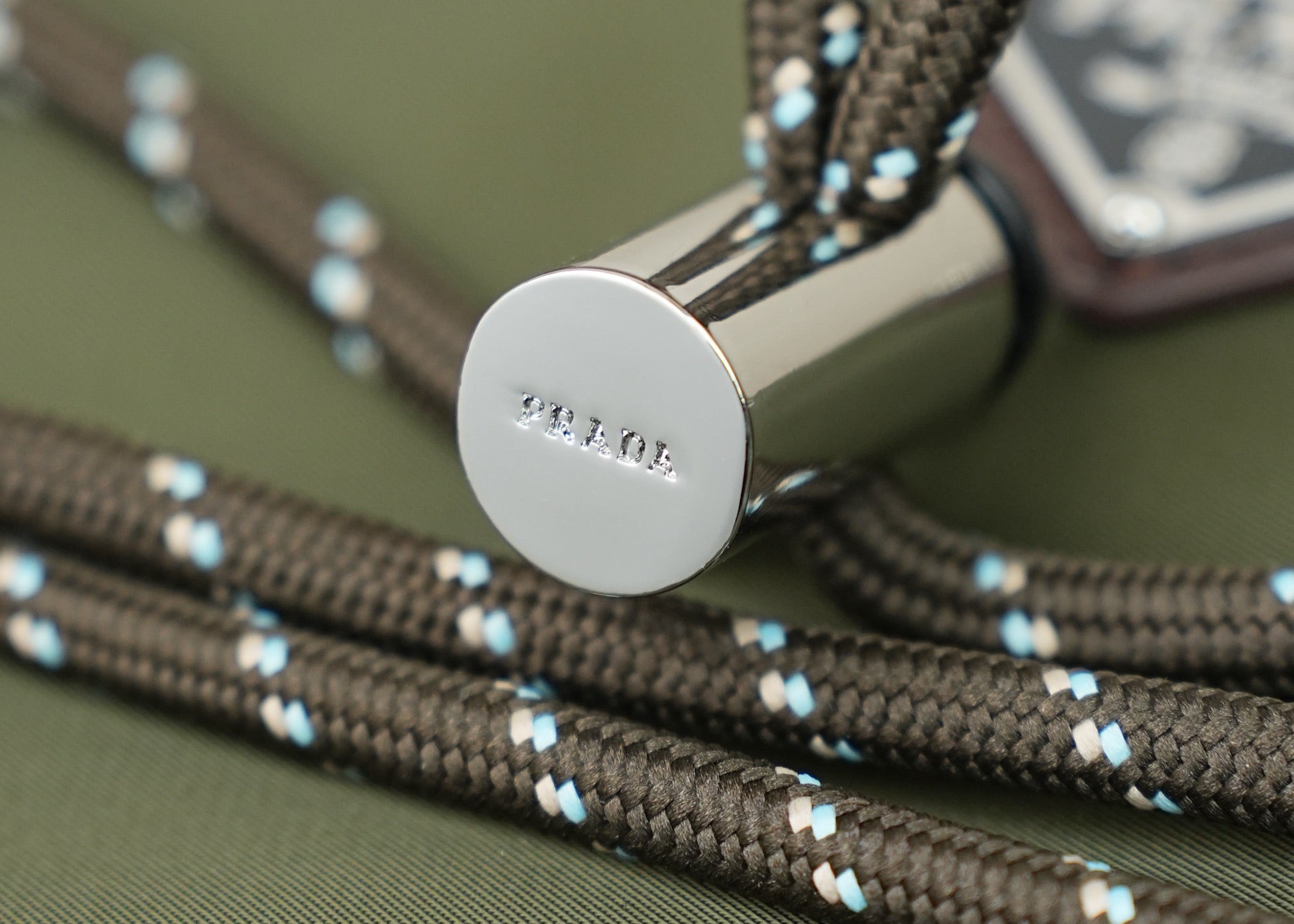 Round silver snap button on green bag with blue-and-beige woven trim cords.