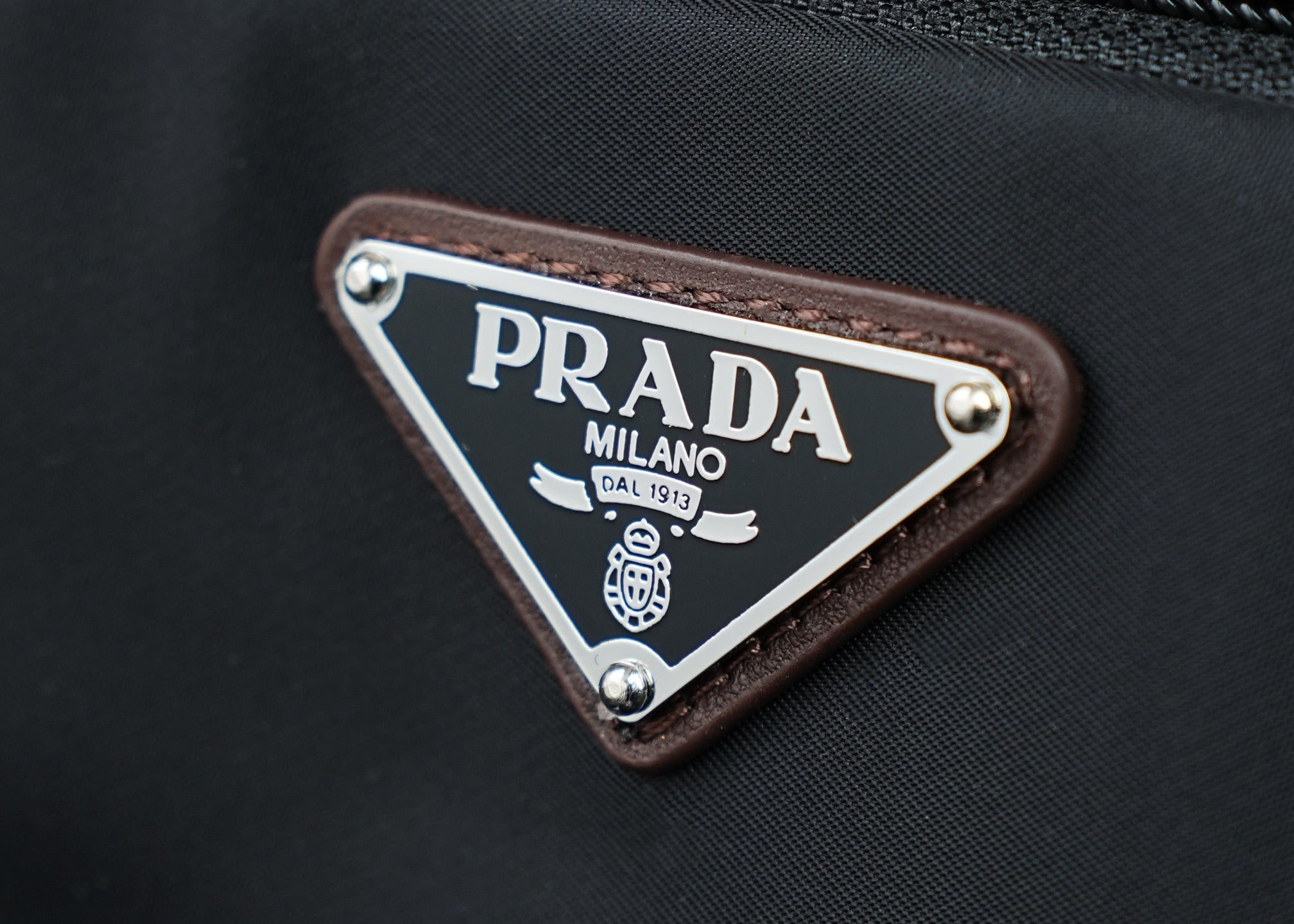 Triangular PRADA logo patch with PRADA MILANO text on brown leather trim.