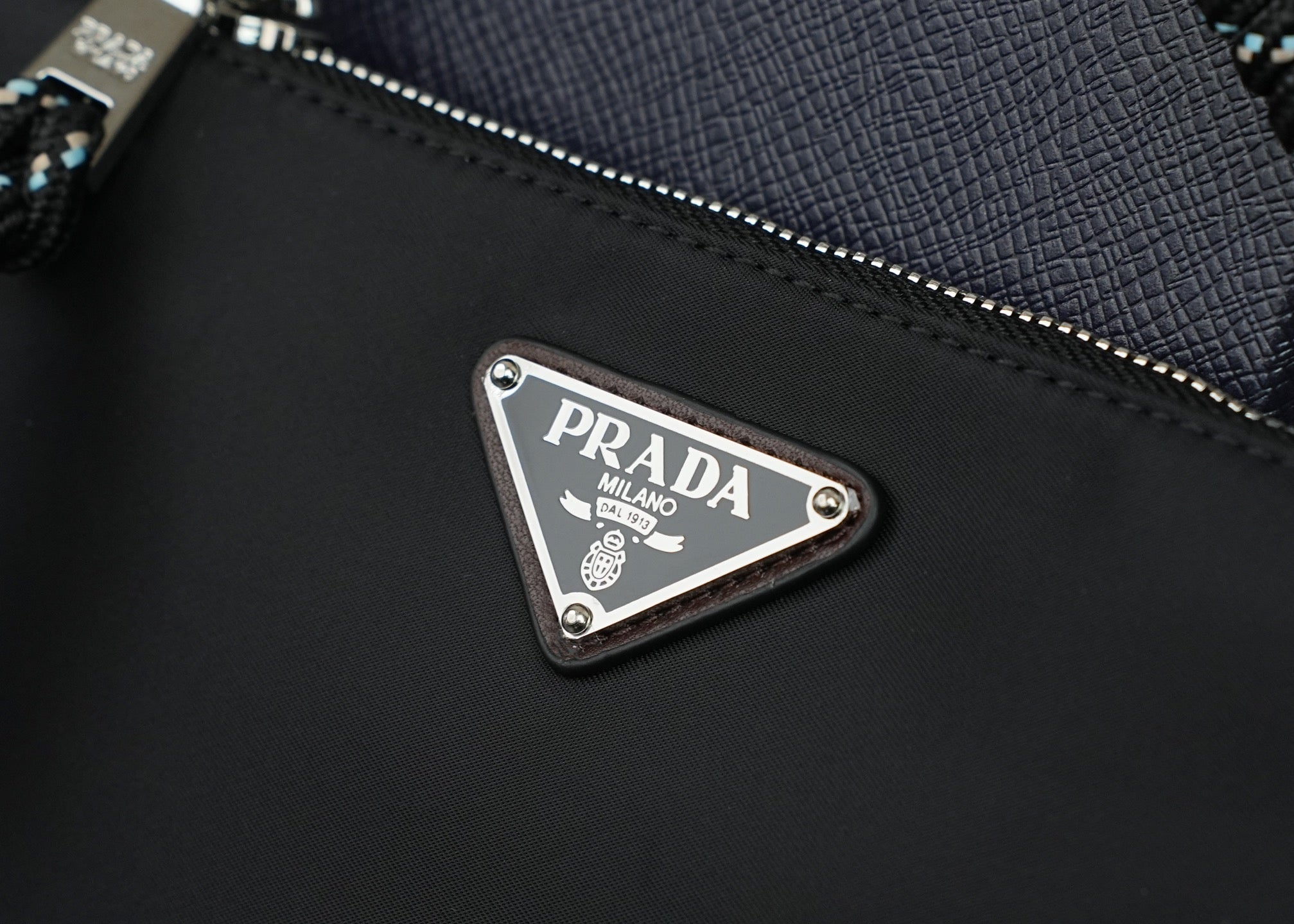 Prada metal logo plate on black textured bag with zipper and stitched edge