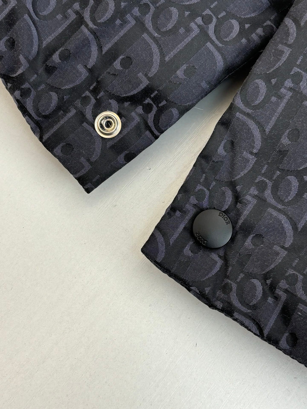 Cuff area of dark Dior monogram vest with round snap button and textured fabric.