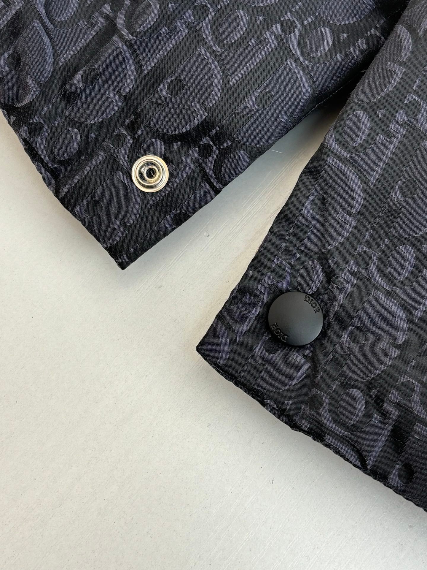 Cuff area of dark Dior monogram vest with round snap button and textured fabric.