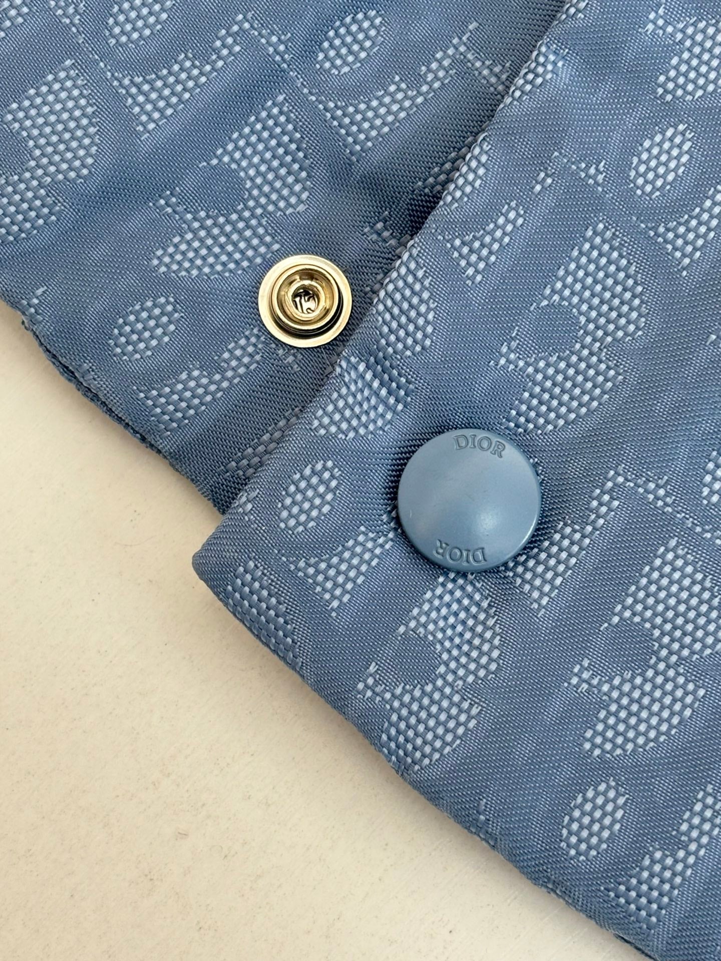Close-up of light blue monogram vest with gold snap button and gray Dior-style logo