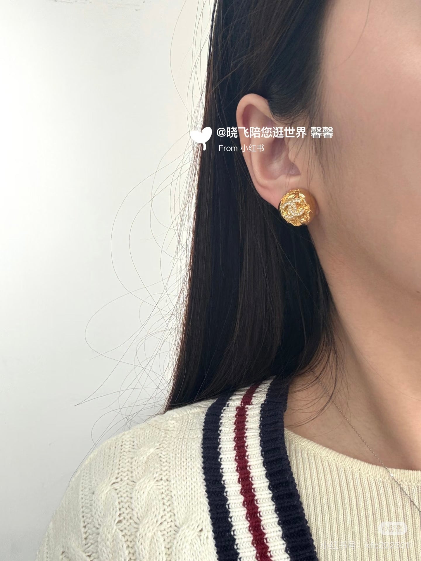 Gold-tone round stud earrings worn on ear with logo and clear rhinestones