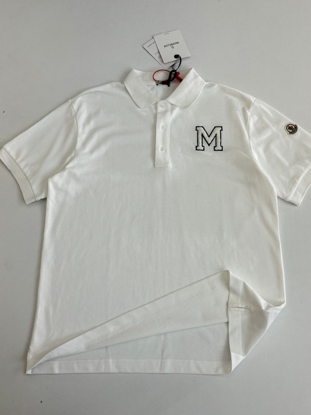 White Moncler polo shirt flat lay with M logo, collar buttons, sleeve studs