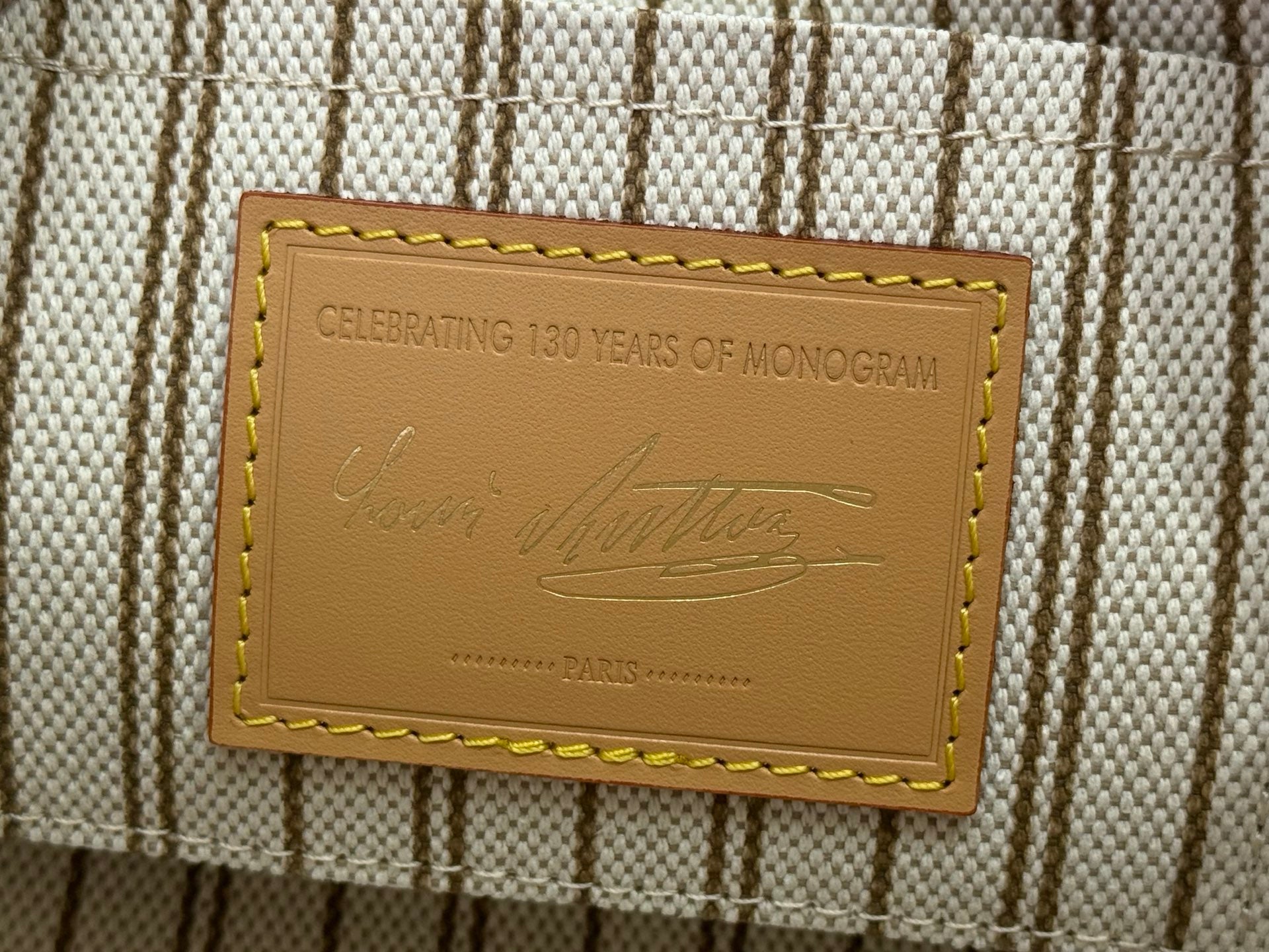 Tan leather label reading “CELEBRATING 130 YEARS OF MONOGRAM” on striped fabric