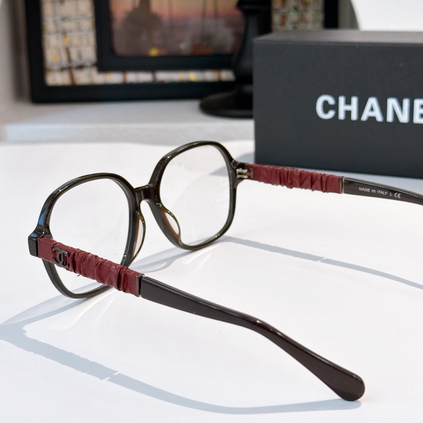 Chanel eyeglasses side view with black frame, burgundy strap-style temple, clear lens.