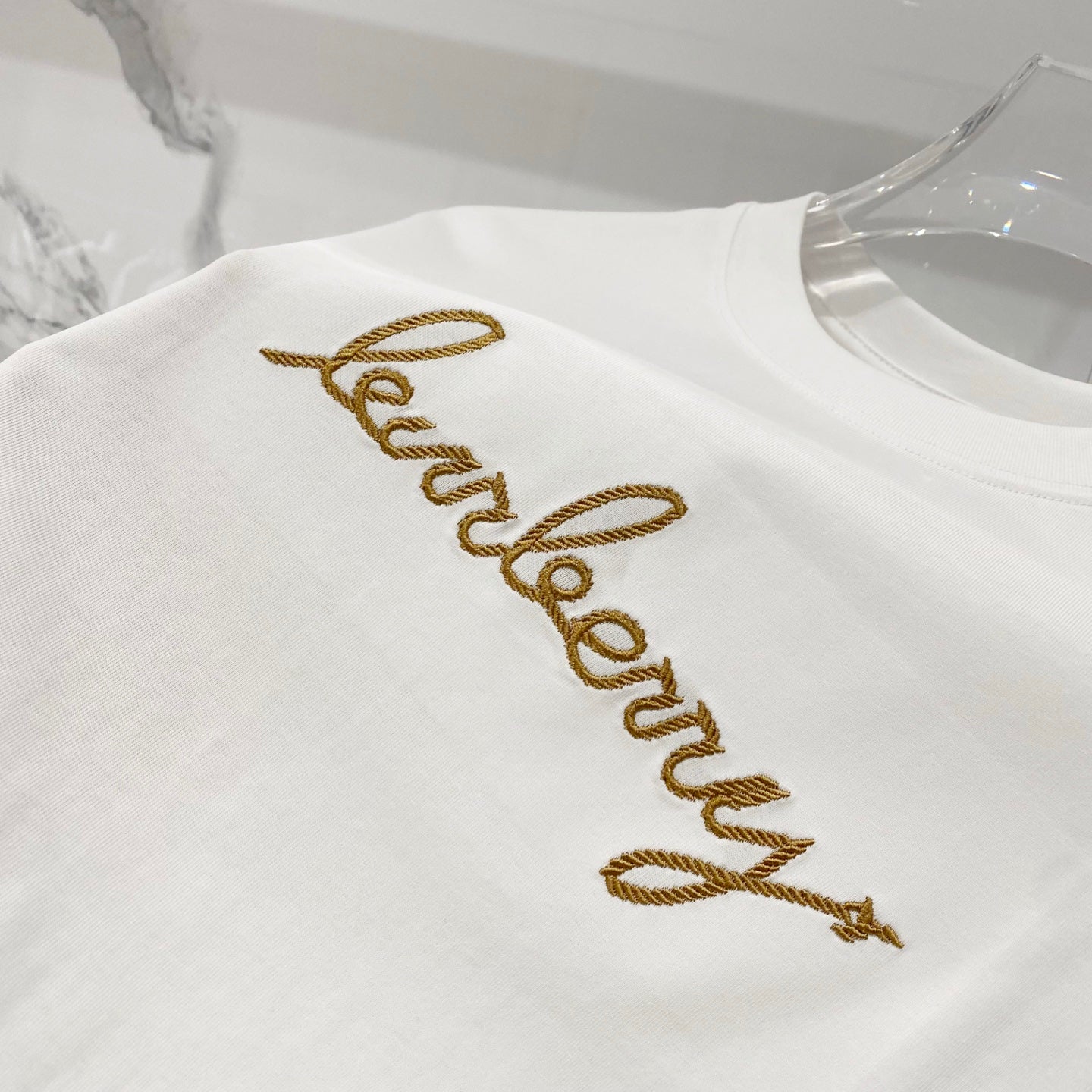 White short-sleeve T-shirt with gold embroidered logo text across the chest