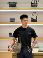 Black short-sleeve T-shirt with tan chest logo patch, worn with hand in pocket