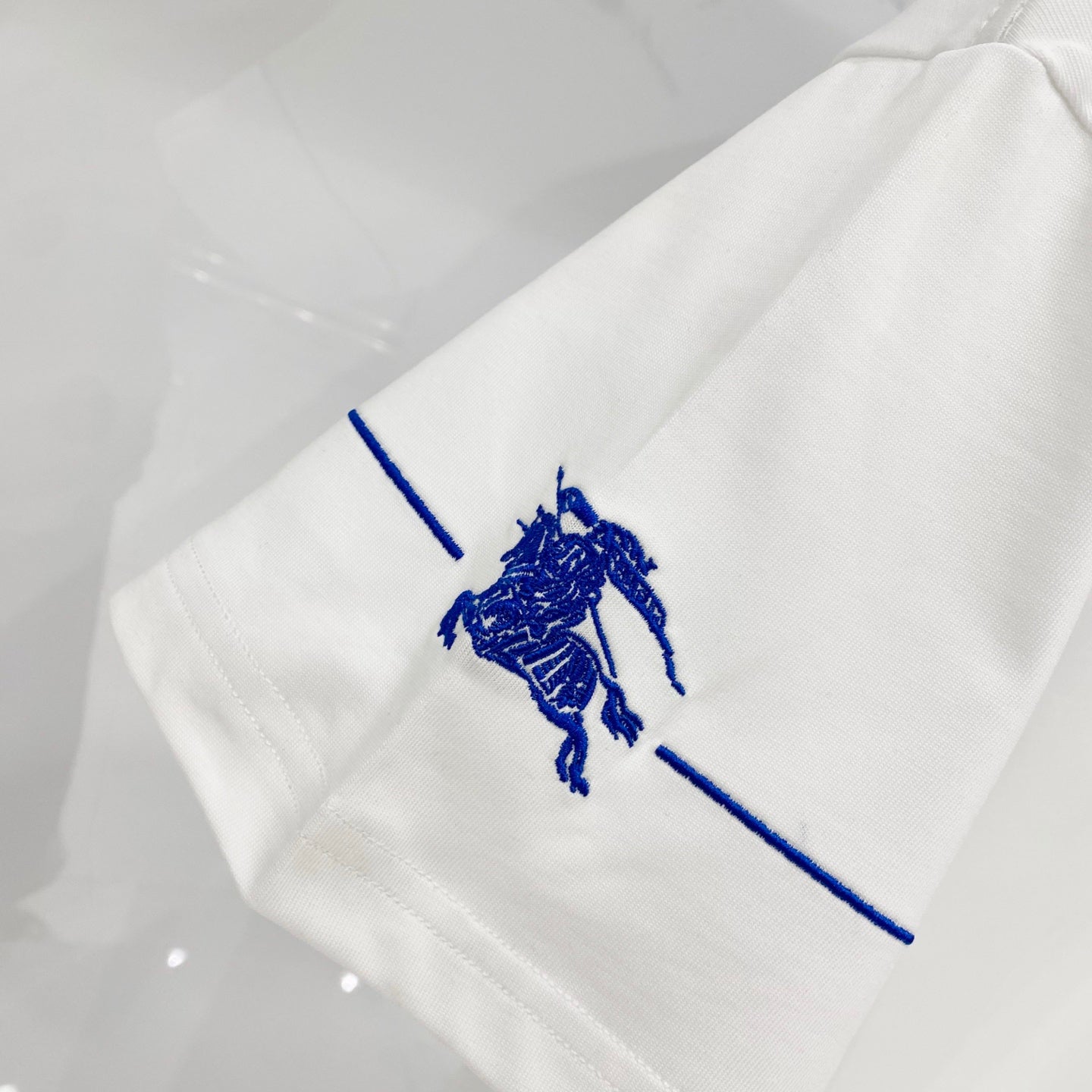 White T-shirt sleeve with small blue horse rider graphic print along the seam.