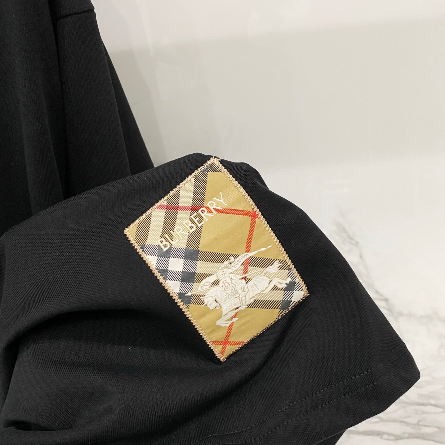 Black T-shirt with multicolor Burberry-style check patch appliqué on the chest.