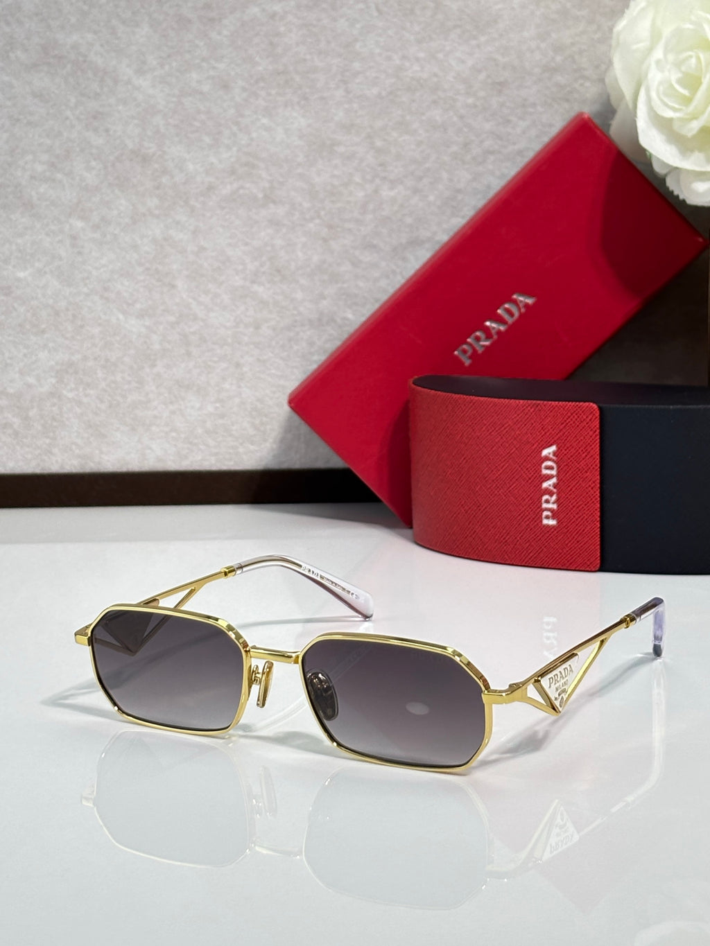 Prada sunglasses with gold frame and dark tinted lenses, shown near a red logo case.