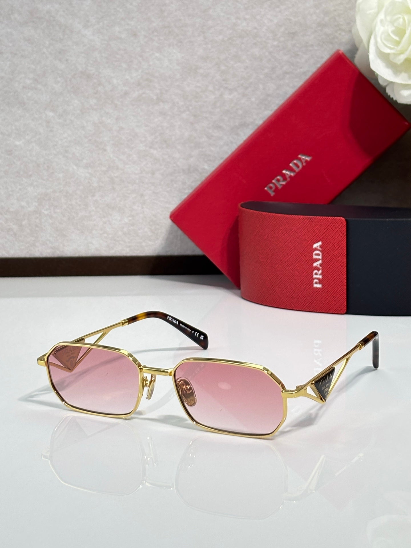 Prada sunglasses with gold frame and pink tinted lenses, beside a red logo case.