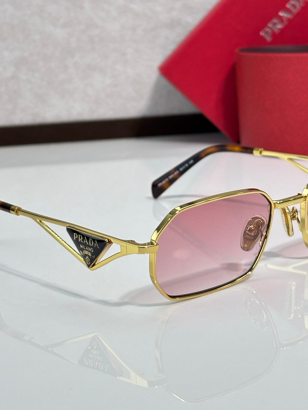 Prada sunglasses close-up, gold frame with pink tinted lens, with red logo case.