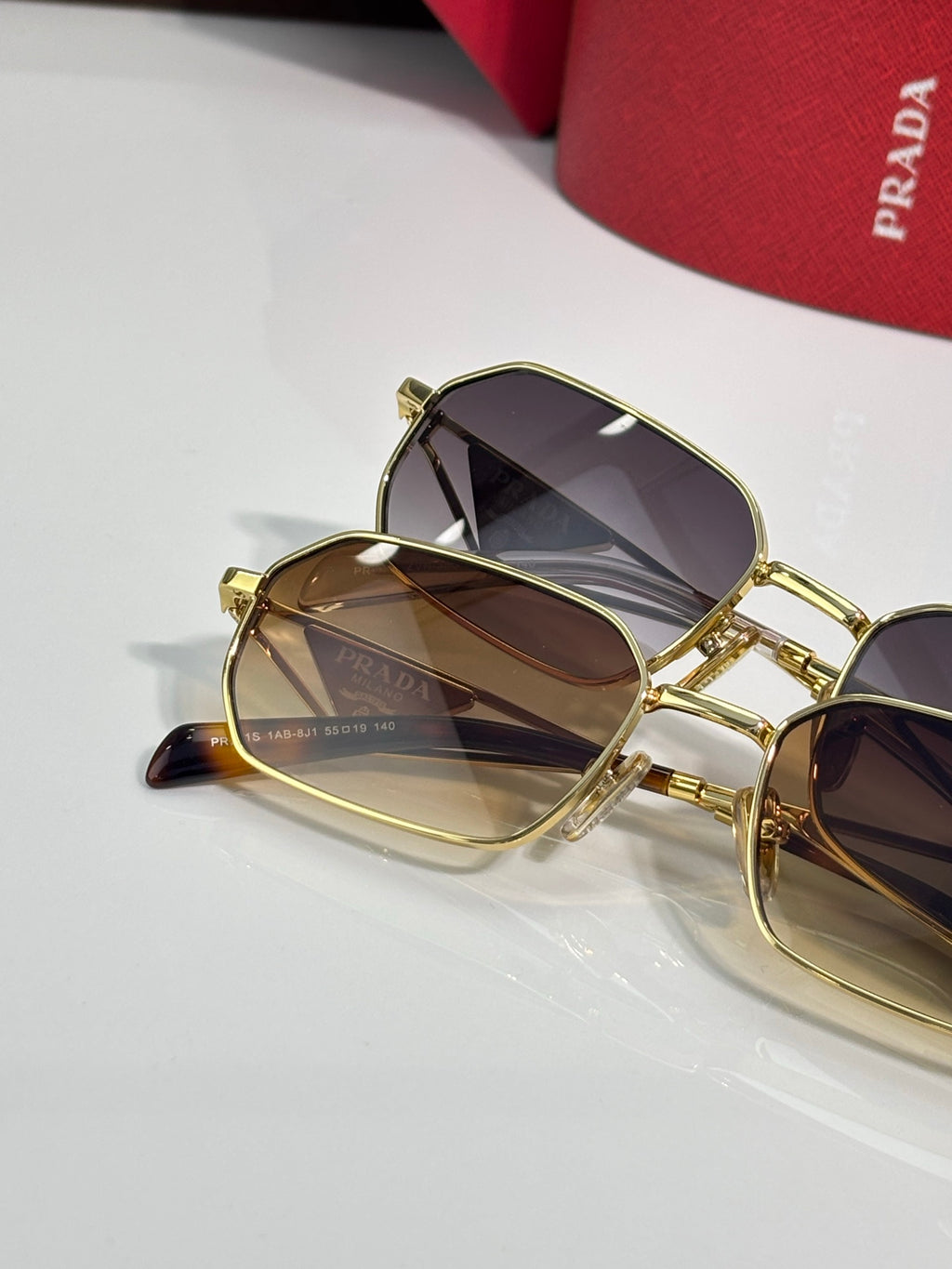 Prada sunglasses close-up, gold frames with dark lenses, beside a red logo case.