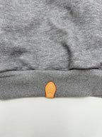 Gray hoodie hem with small orange leather-look logo patch at the center