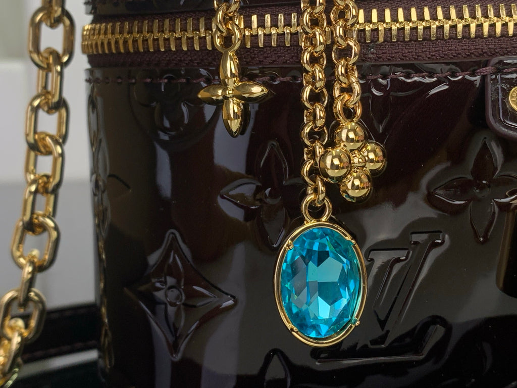 Blue oval gemstone charm on gold chain attached to glossy monogram handbag