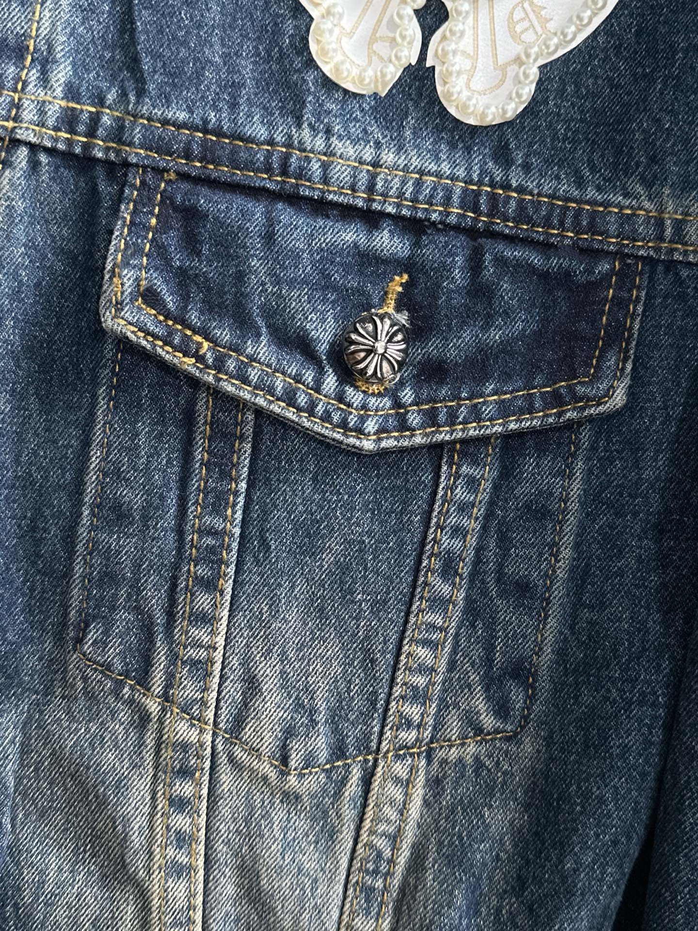 Blue denim jacket front pocket with small gold-tone button and central emblem pin