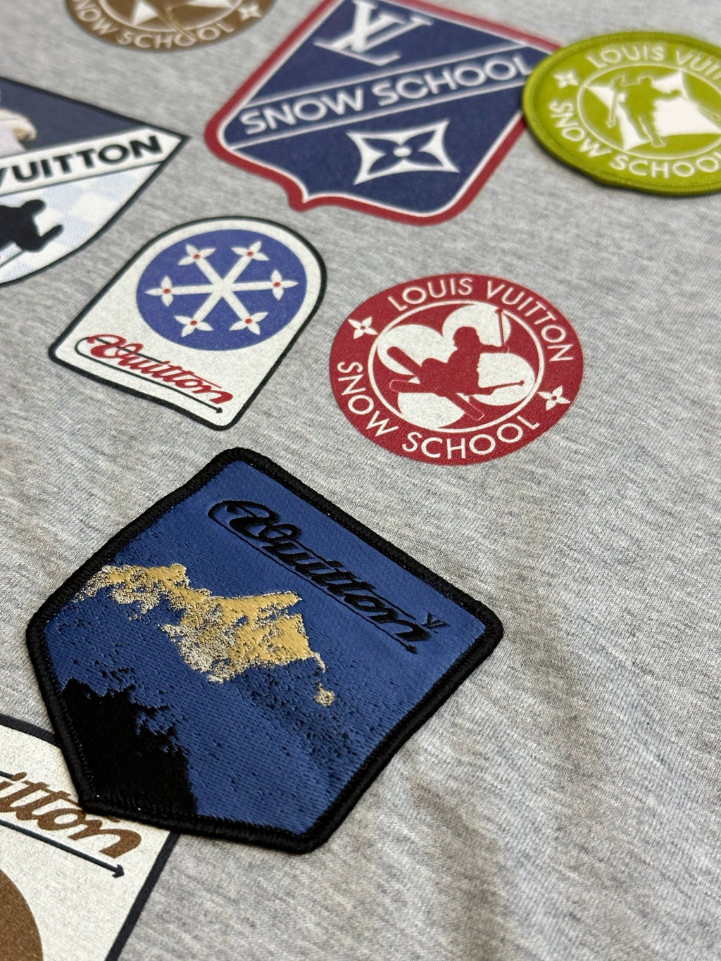 Grey T-shirt with varied logo patches, including a blue rectangular patch