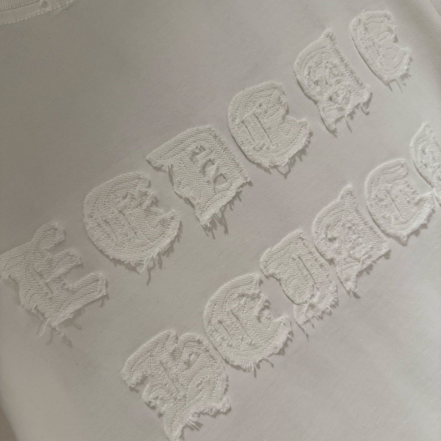 Raised embroidered logo detail on white T-shirt, showing textured stitching pattern.