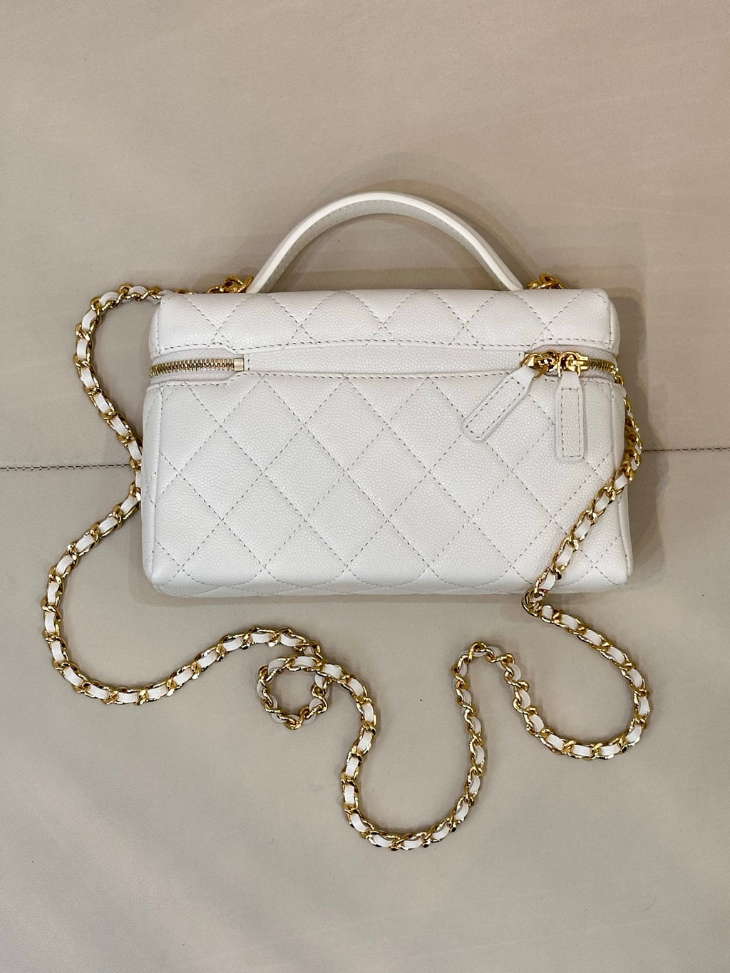 White quilted leather handbag with top handle, front zip pocket, and gold chain strap.