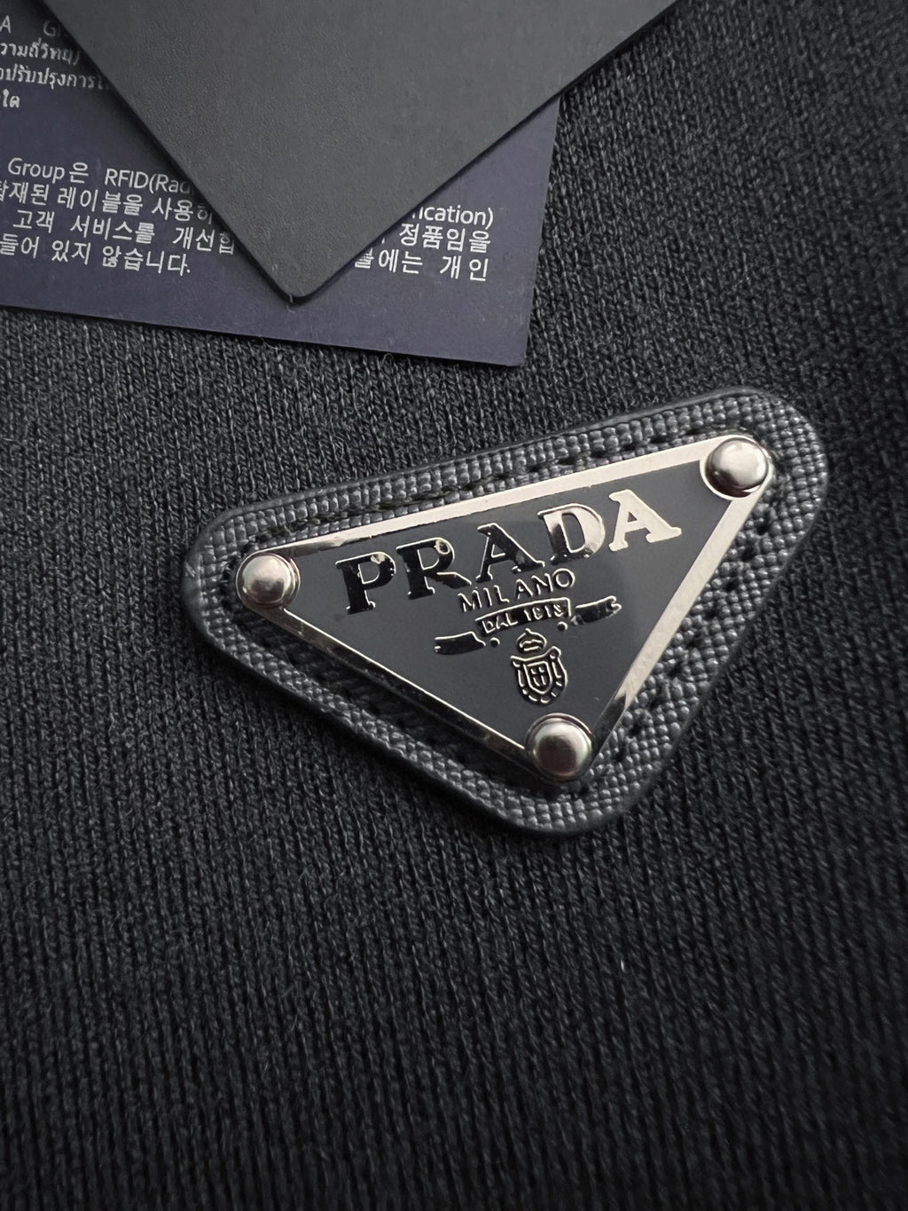 Prada woven logo patch on black fabric, triangular metal-trimmed emblem
