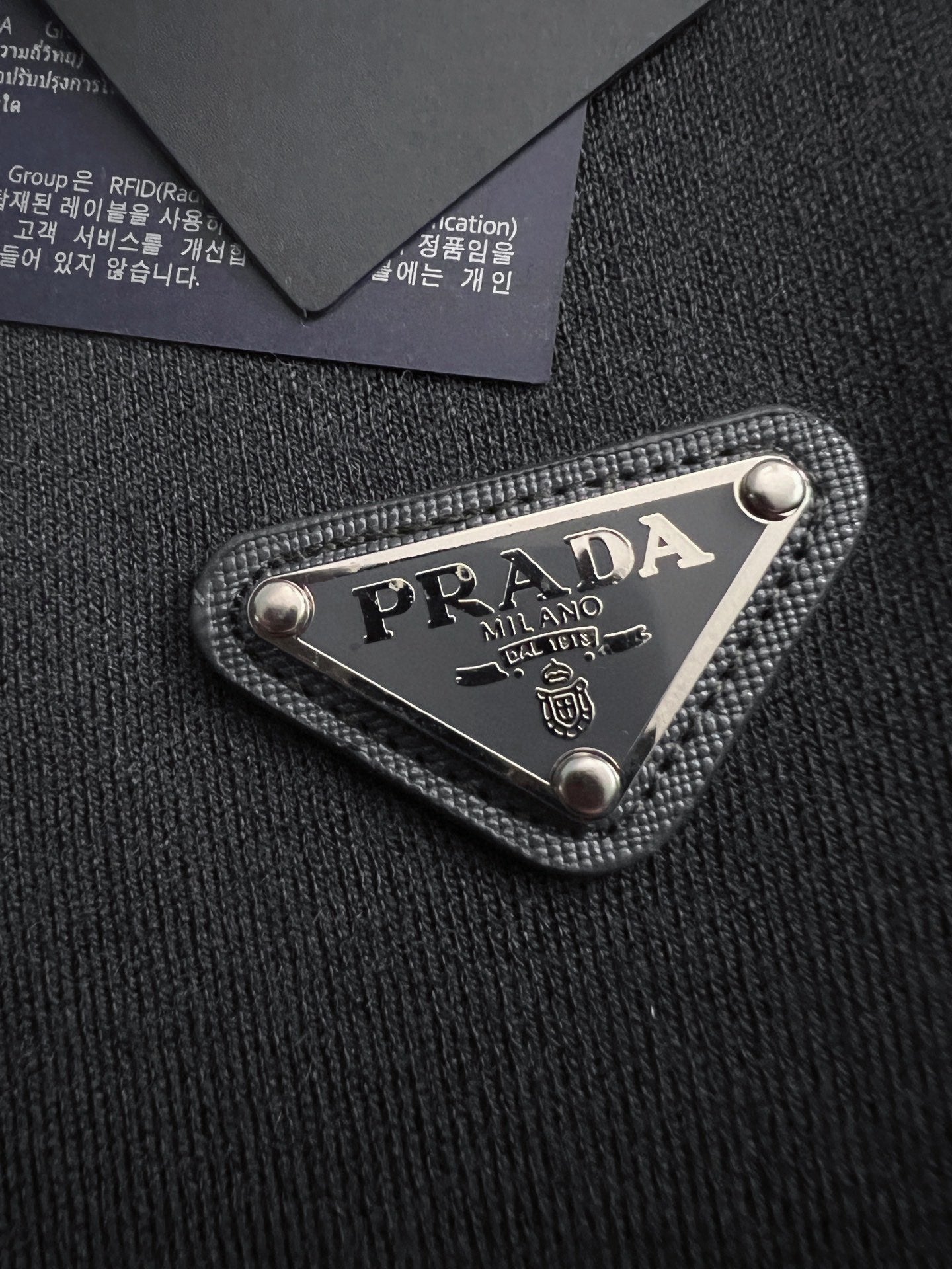 Prada woven logo patch on black fabric, triangular metal-trimmed emblem