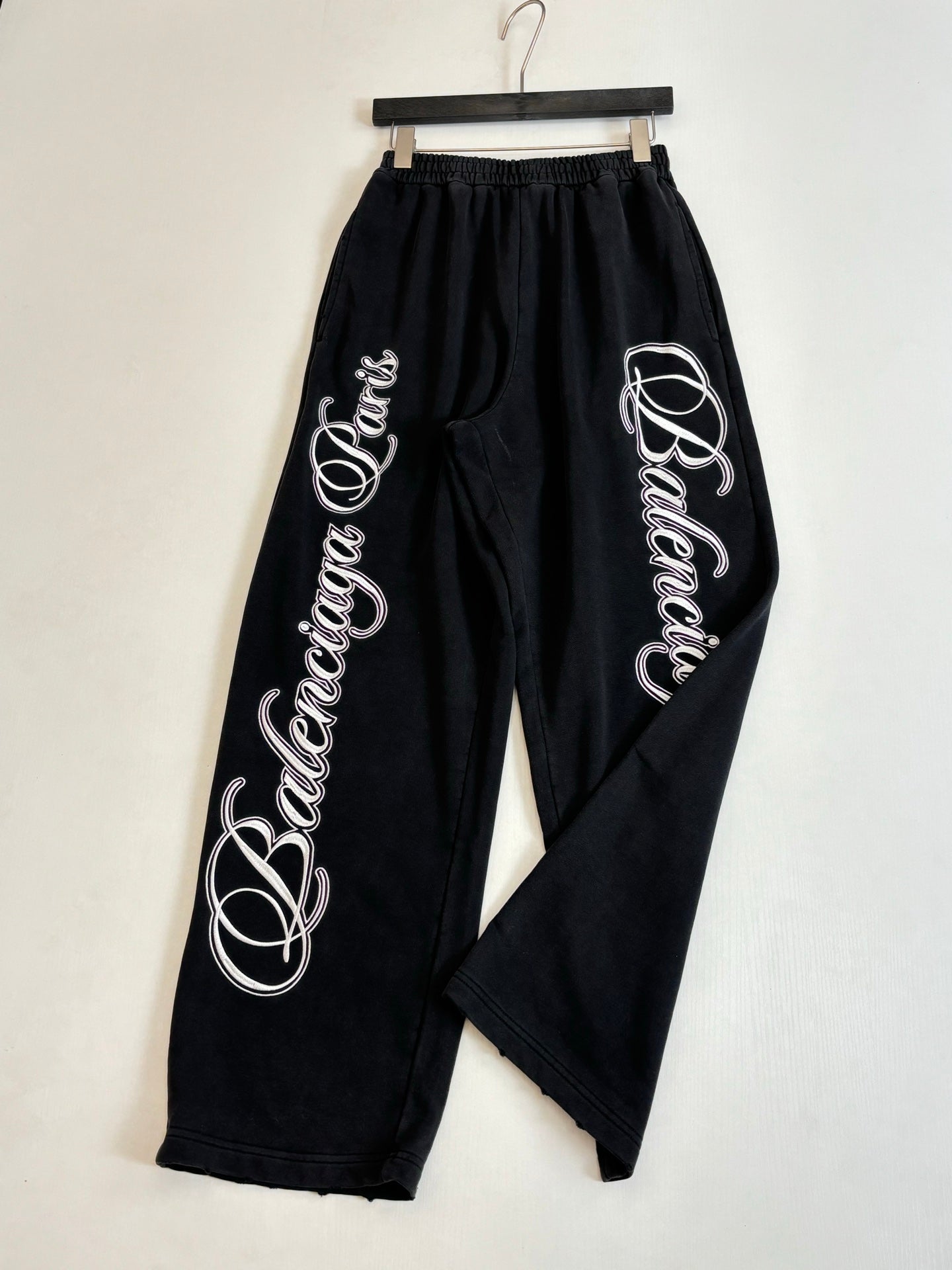 Black Balenciaga track pants with large side script logos, hanging on hanger