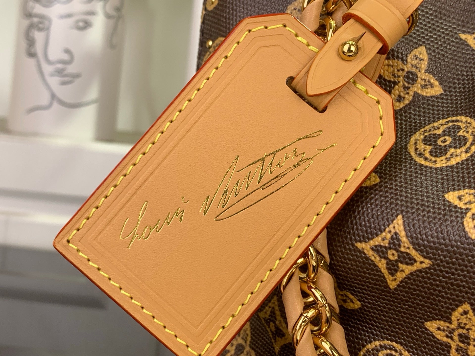 Tan leather luggage tag with stitched edge and cursive-style logo on monogram canvas