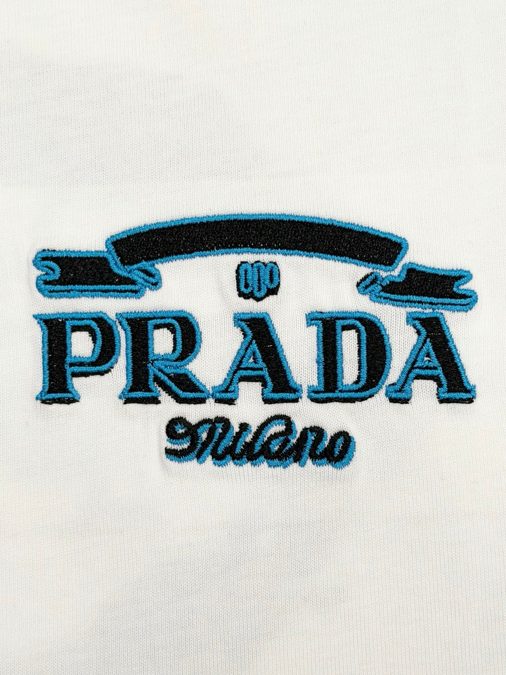 Close-up of embroidered PRADA logo with blue stylized text on white T-shirt