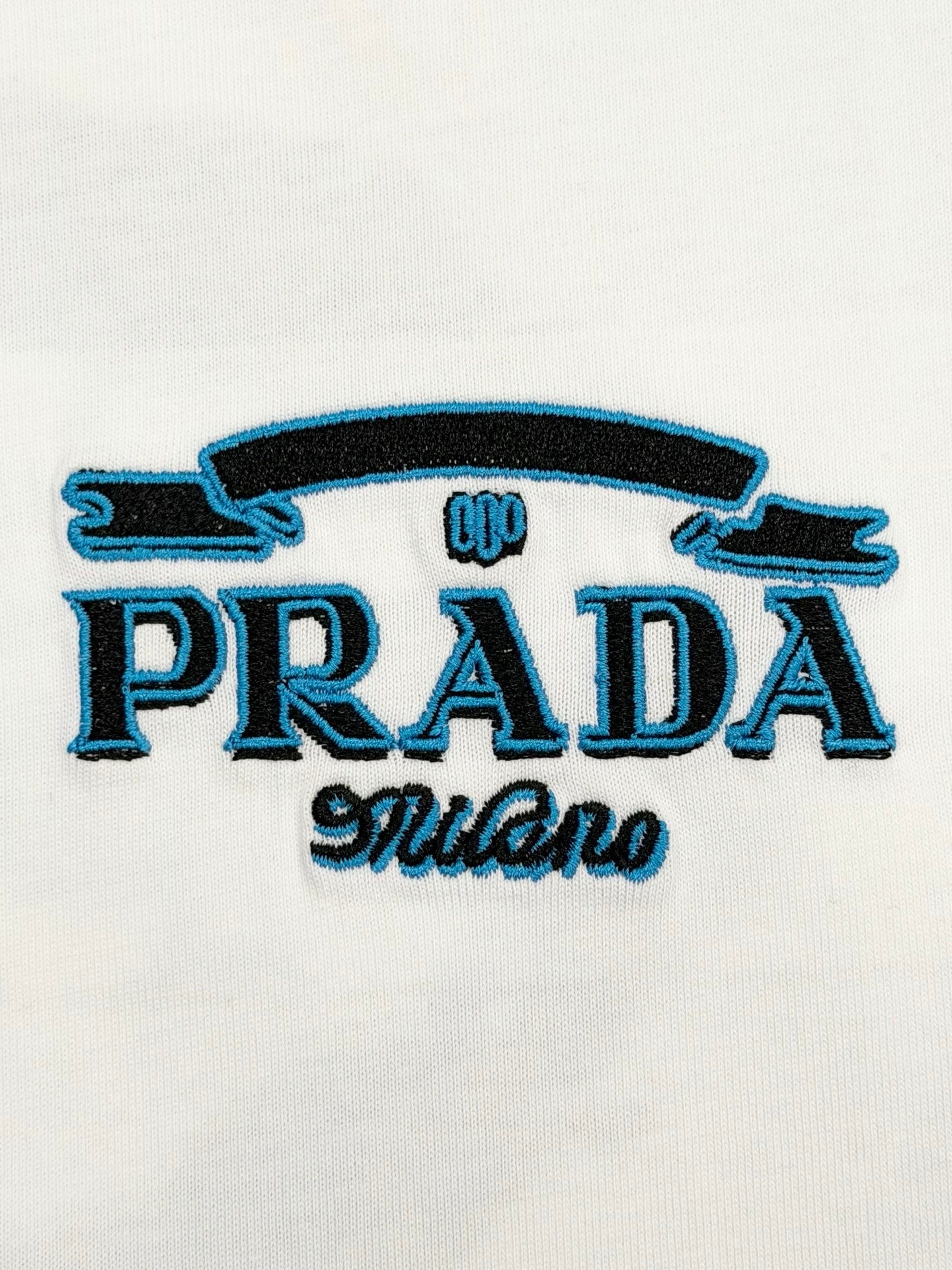 Close-up of embroidered PRADA logo with blue stylized text on white T-shirt
