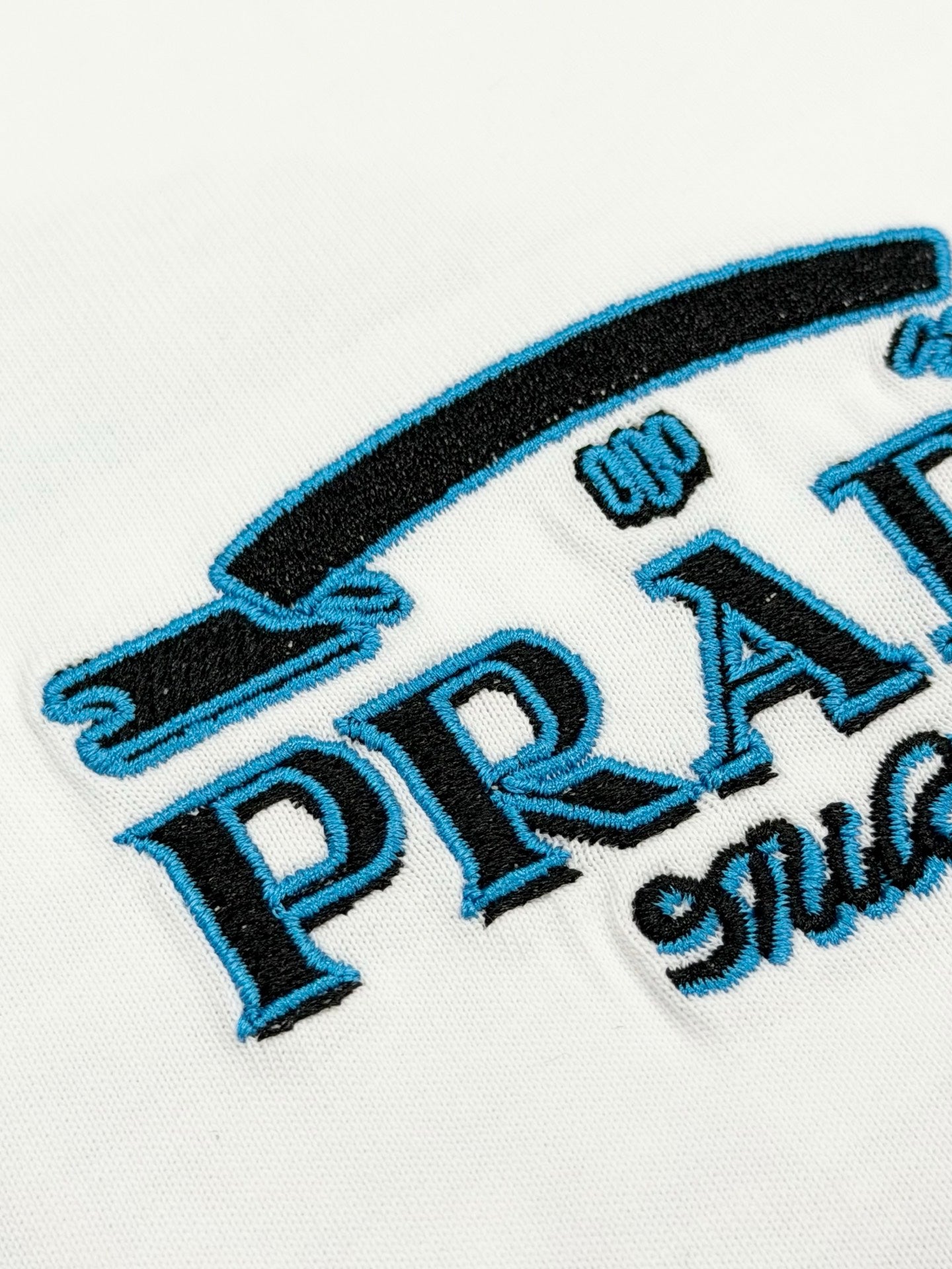 Close-up of embroidered PRADA logo and blue lettering on white T-shirt