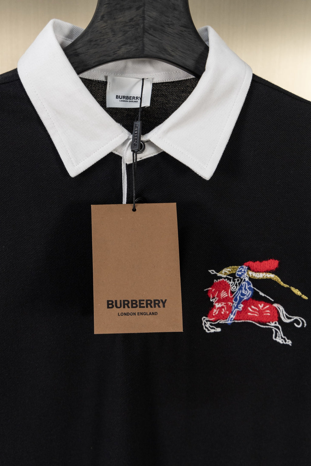 Black polo shirt with white collar, embroidered knight-and-horse logo, and hanging tag