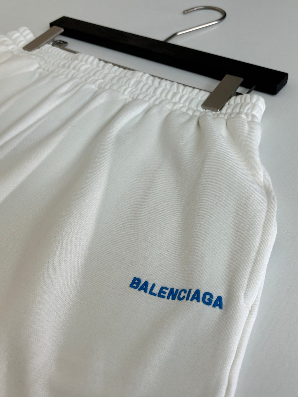 White Balenciaga shorts close-up with elastic waistband and blue BALENCIAGA logo print.
