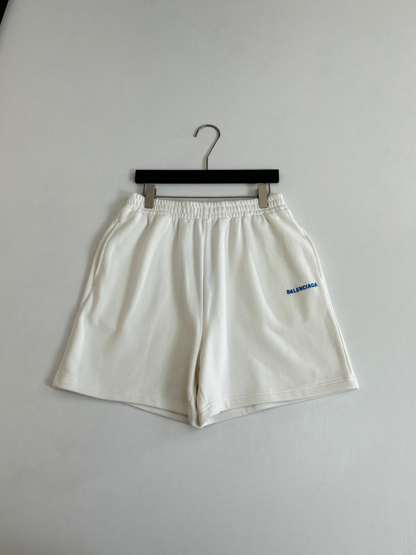 White Balenciaga shorts on hanger with elastic waistband and blue logo on leg.