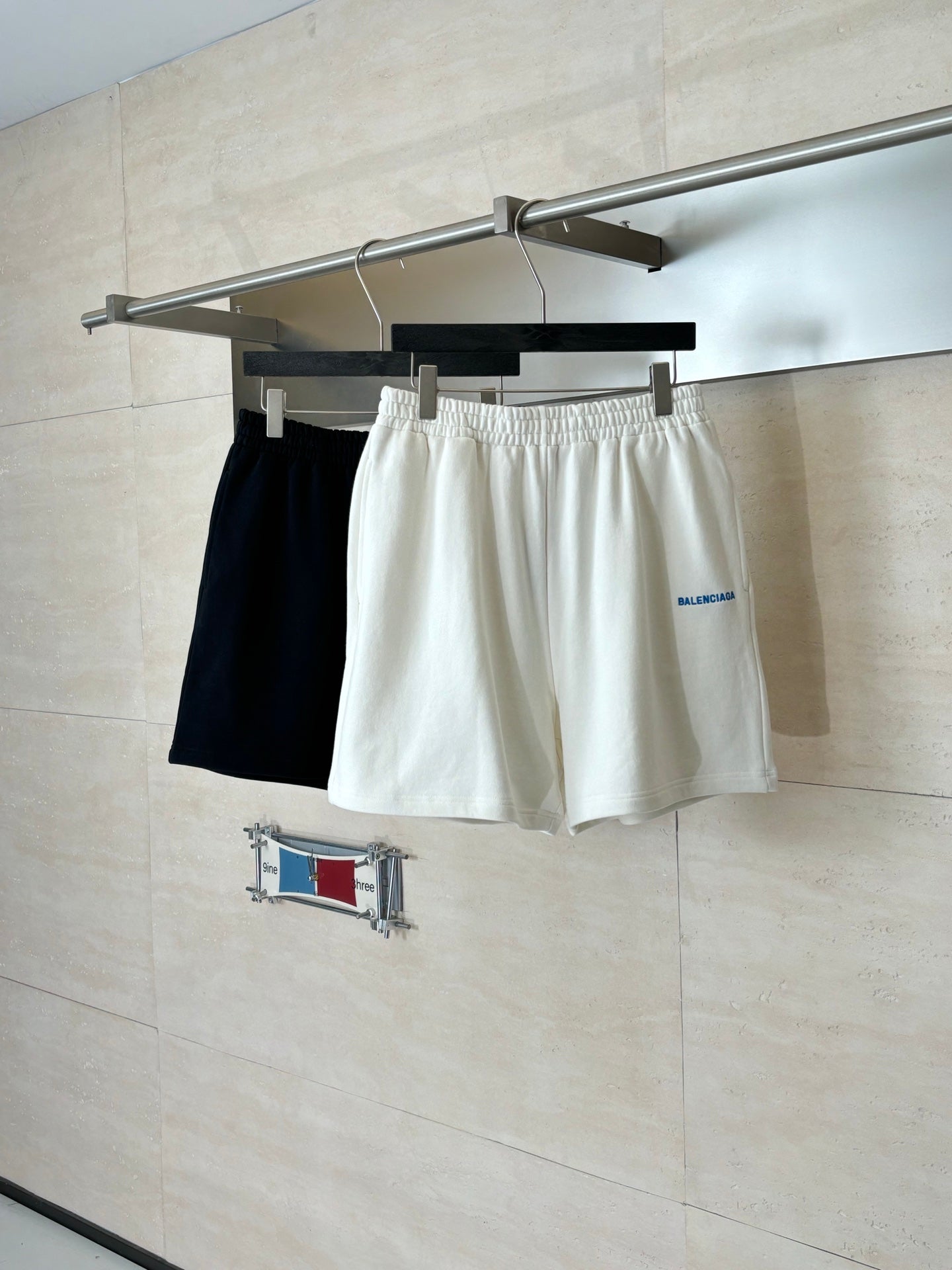White Balenciaga shorts on hanger with elastic waistband; dark shorts partially visible behind.
