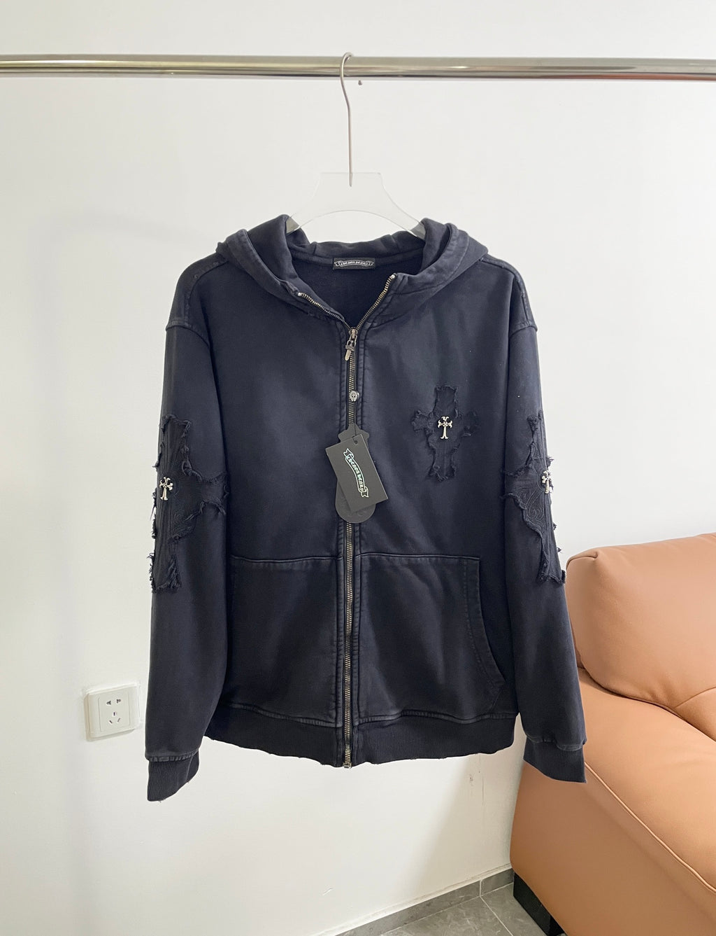 Dark zip-up hoodie with hood, front pockets, and cross emblem on chest and sleeves