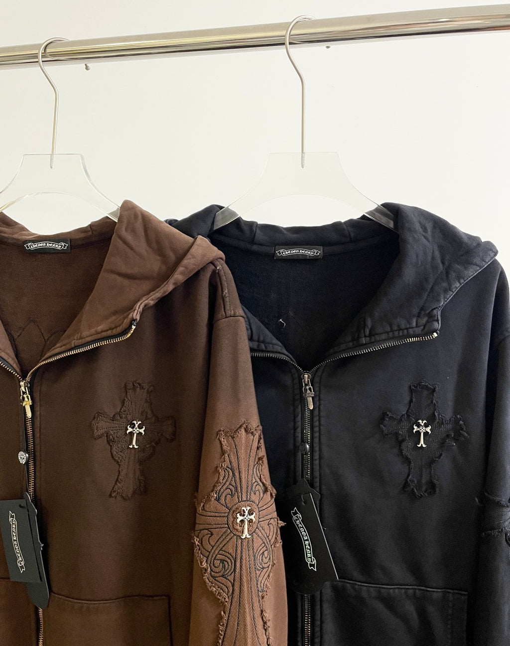 Brown and black zip-up hoodies side-by-side on hangers, embroidered cross logos
