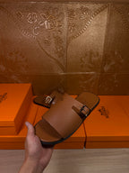 Brown leather slide sandal held in hand, with silver buckle and wide upper strap