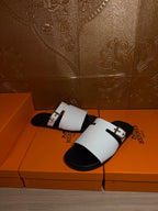 Hermes sandals with white slide straps and silver buckles, diagonal view on orange boxes