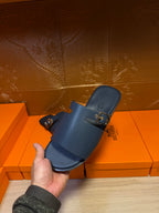 Hermes slide sandals held up, dark blue leather slide with two gold buckled straps.