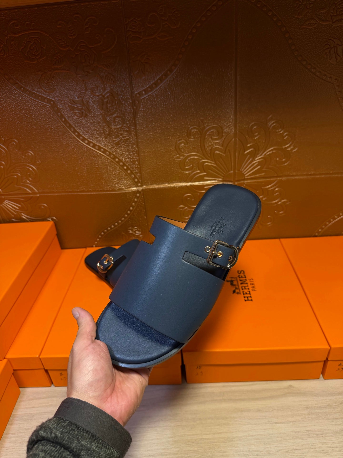 Hermes slide sandals held up, dark blue leather slide with two gold buckled straps.