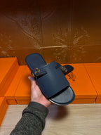 Hermes slide sandals held in hand, dark blue leather with gold buckles and wide strap.