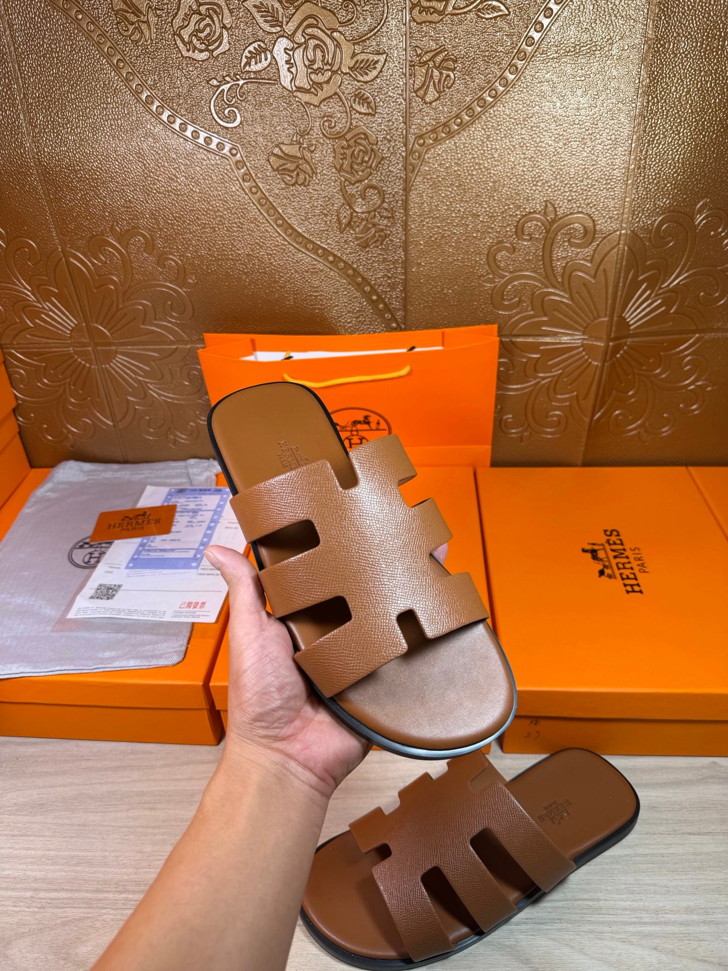 Brown slide sandals held in hand; wide cutout straps and textured footbed with flat sole.